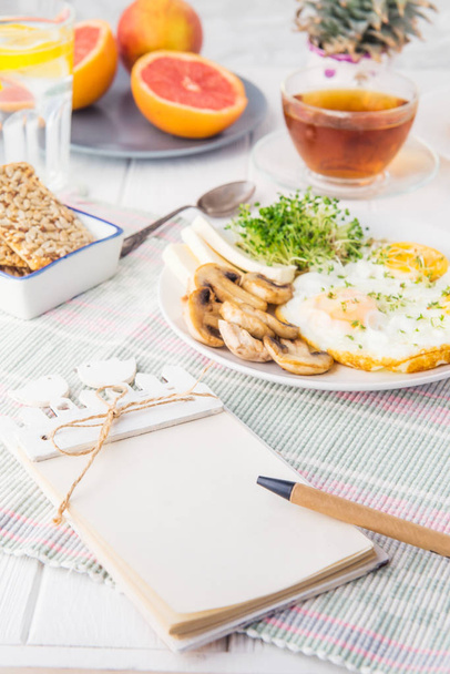Notebook with emty blanks and healthy breakfast plate with scrambled eggs, cheese, grilled mushrooms and sprout micro greens on the served white wooden table. Selective focus. - Φωτογραφία, εικόνα