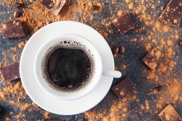 Tasse schwarzer Kaffee auf dunklem Steinhintergrund mit Bohnen, Kakaopulver und rundherum verstreuten Schokoladenstücken. Selektiver Fokus. Raum für Text. - Foto, Bild