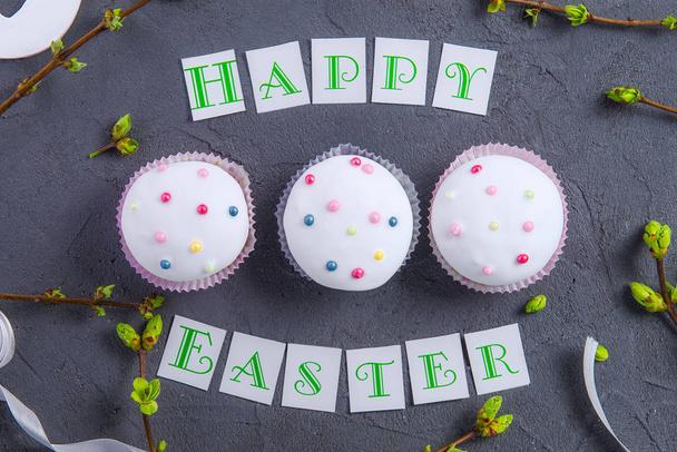 Top view holiday composition of Happy Easter lettering, branches with young shoots of greenery and three decorated cupcakes on dark stone background. Art concept. Selective focus. - Foto, immagini
