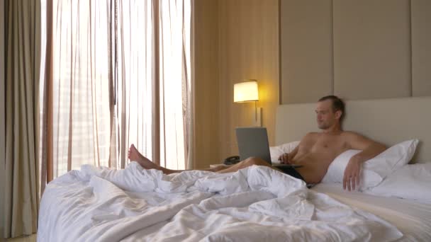Νεαρός κάθεται στο κρεβάτι με τον φορητό υπολογιστή ανοιχτά και το smartphone. Hes που εργάζονται σε αυτό. 4k - Πλάνα, βίντεο