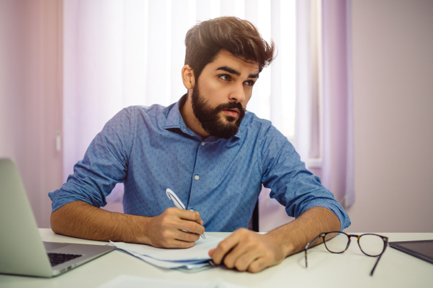 Νέος όμορφος αρσενικό δημοσιογράφος που εργάζονται με έγγραφα σε χαρτί και το laptop. - Φωτογραφία, εικόνα