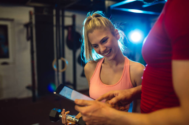 Smiling personal trainer using digital tablet while talking to blonde woman at gym. Happy couple using tablet in fitness club. Client trainer looking at computer her progress at the gym. - Photo, Image