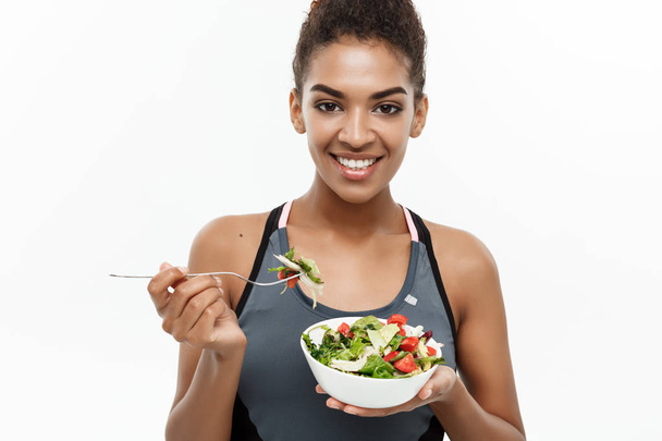 Terve ja Fitness-konsepti Kaunis amerikkalainen afrikkalainen nainen kuntoiluvaatteissa ruokavaliossa syö tuoretta salaattia. Eristetty valkoisella pohjalla
. - Valokuva, kuva