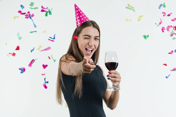 Positive smiling woman at celebration party with a glass of wine and in a festival hat, Point your finger at you - 写真・画像
