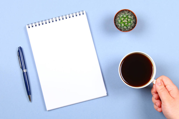 Empty notebook with pen, coffee and cactus on a blue background. Copy the space. Top view. A hand holds a cup of coffee. - Foto, Bild