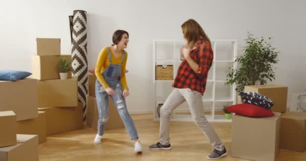 Young joyful just married couple dancing, having fun and fighting with pillows among unpacked boxes while moving in the new modern apartment. Indoors - Footage, Video