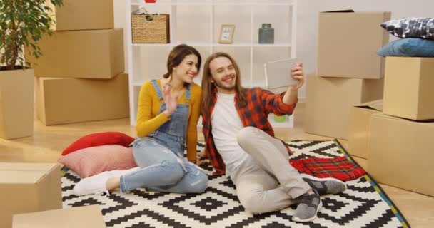 Jonge mooie paar zittend op het tapijt op de vloer onder de uitgepakte vakken, videochat hebben en tonen van hun nieuwe appartement. Binnenshuis - Video