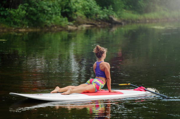 Young athletic woman doing fitness on a board with an oar on a lake - Photo, Image