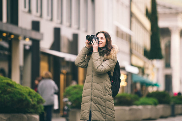 Beautiful young tourist woman stands on the background of the central street in Munich in Germany in winter. Holds a black big professional camera, takes a photo and smiles - Fotoğraf, Görsel