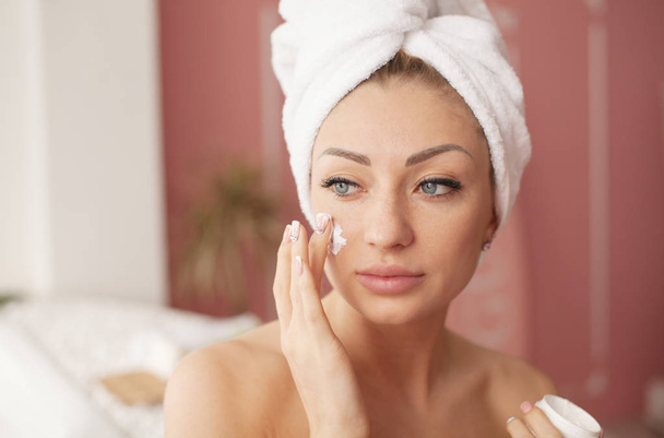 Mulher jovem com pele impecável, aplicando creme hidratante em seu rosto. Foto da mulher após o banho em roupão branco e toalha. Conceito de cuidados da pele
 - Foto, Imagem