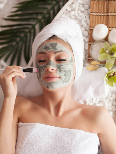 Beautiful woman having clay facial mask apply by beautician. - Zdjęcie, obraz