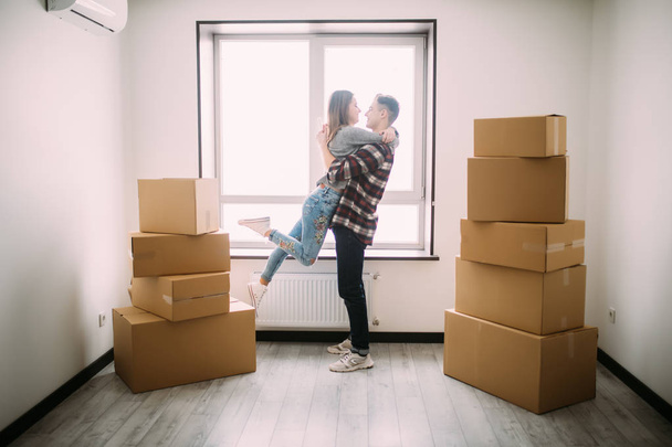 Full length portrait of beautiful young couple hugging and smiling while standing among moving cardboard boxes in empty room - Fotografie, Obrázek
