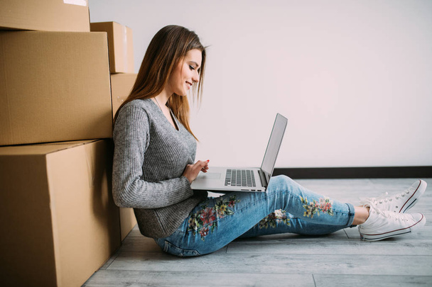 Happy young woman moving to new home sitting on the floor and use laptop near boxes - Photo, Image