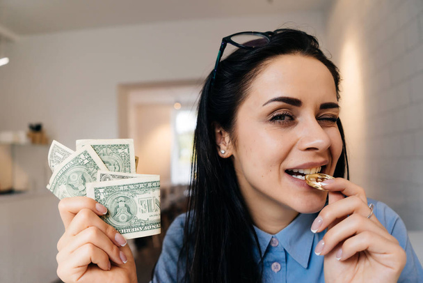 attractive young girl in blue shirt holds dollars and bites golden bitcoin - Zdjęcie, obraz
