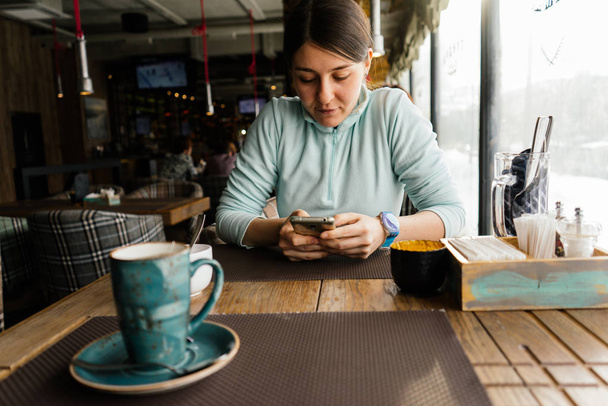 cute young girl in a blue sweater sits in a cafe and waits for her food, looks into a smartphone - Photo, Image