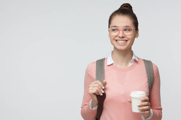 Indoor photo of beautiful young female in pink eyeglasses isolated on grey background with bag on her back, smiling, holding a cup of coffee to go. Copyspace - Foto, immagini