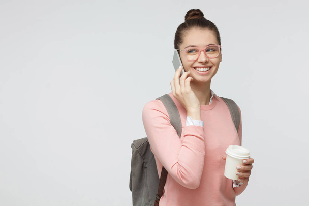 Studio photo of student girl isolated on grey background with backpack on her back, holding takeaway coffee cup while talking on smartphone, smiling positively and looking sideways  - Foto, Imagem