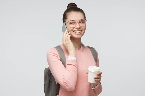 Side portrait of young European female isolated on gray background wearing pink eyeglasses, listening to smartphone, showing satisfied happy smile, holding coffee to go cup - Fotó, kép
