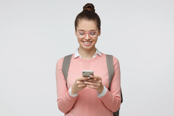 Indoor shot of good looking female student isolated on gray background looking at smartphone, smiling openly while holding smartphone in both hands - Foto, Imagen