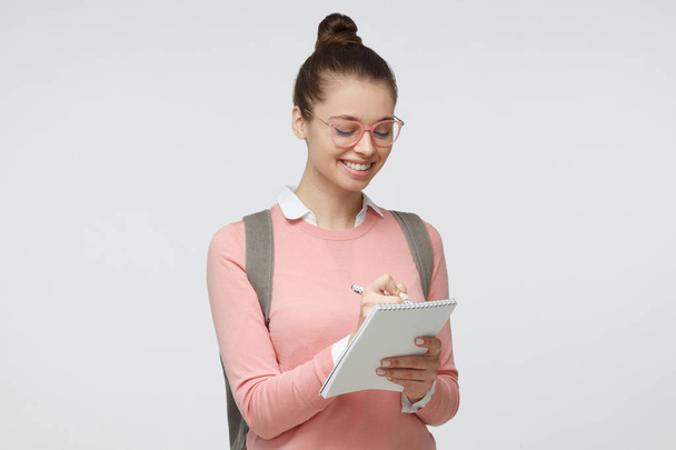 Indoor shot of young female in glasses against grey background taking notes on blank sheets of writing pad she is holding with satisfied and interested smile, happy to study - Fotografie, Obrázek