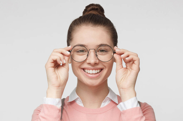 Closeup of attractive young woman isolated on gray background, wearing big round spectacles with thin black frame, touching temples with fingers, smiling - Фото, изображение