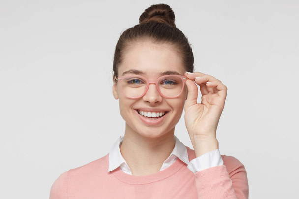 Indoor portrait of young good looking girl in trendy pink glasses isolated on gray background wearing brown hair in bun, smiling, expressing happiness, joy and satisfaction with life - Foto, Bild
