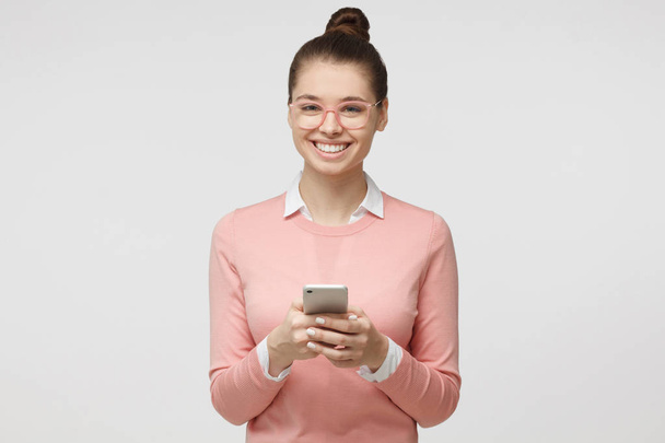 Indoor shot of good looking young woman isolated on gray background looking at smartphone, smiling openly while holding smartphone in both hands - Foto, Imagen