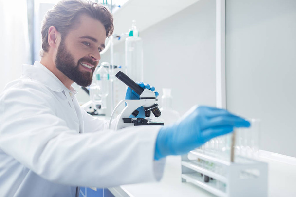 Biological research. Positive smart male biologist holding a microscope and taking a test tube while conducting a biological research - Photo, Image