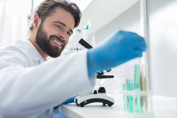 Biologický výzkum. Veselý chytrý mužský vědec, který se dívá na testovací trubky a usmívá se při práci v bio laboratoři - Fotografie, Obrázek