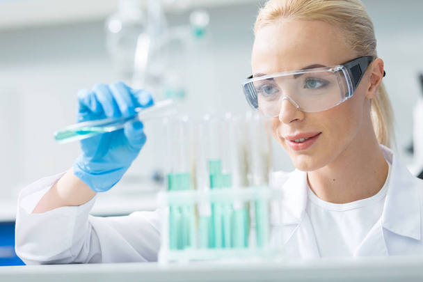 Modern kimya. Neşeli güzel kadın kimyager koruyucu gözlük giyen ve bir deney yaparken test tüpleri bakarak - Fotoğraf, Görsel