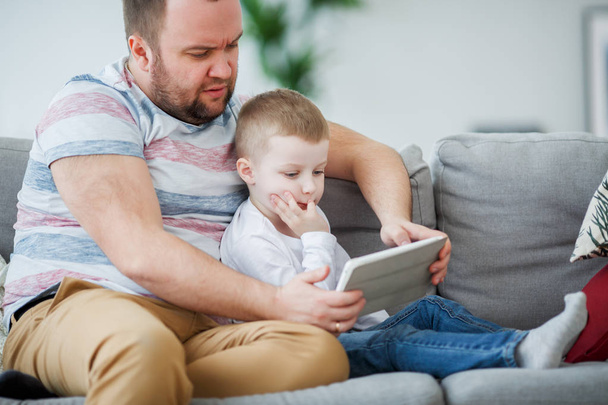 Imagen del padre con el hijo con la tableta sentada en un sofá gris
 - Foto, Imagen