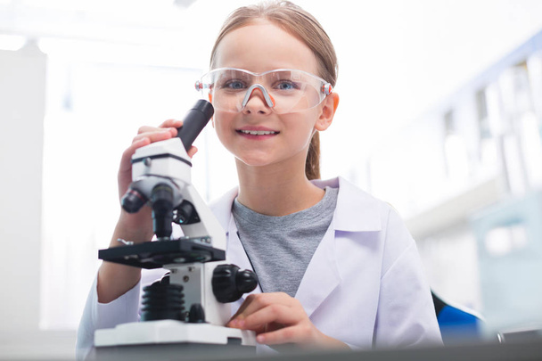 Observation process. Low angle of blond beautiful little girl staring at camera while tuning microscope and wearing lab coat - Foto, Imagen