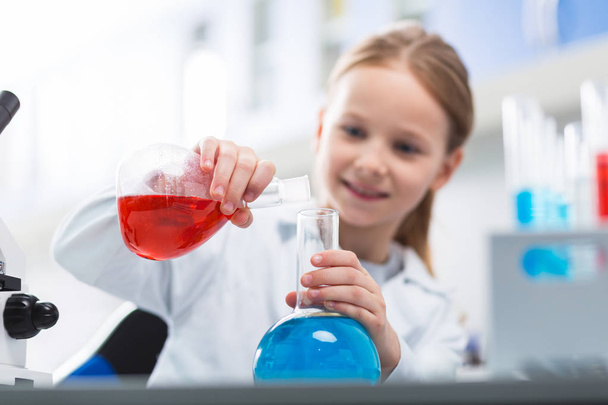 Interesting experiment. Selective focus of glass transparent bulbs hold by little girl who staring at them and dressing in lab coat - Foto, Bild