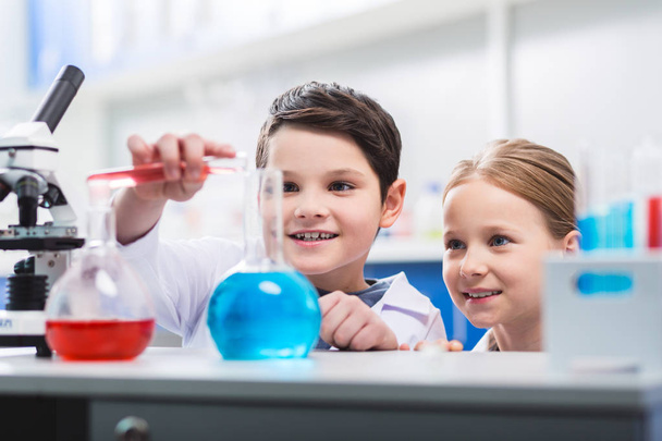 Roekeloos gedrag. Vrolijke vrolijke kinderen staren op laboratorium brillen terwijl ze glimlachend en experiment voeren - Foto, afbeelding