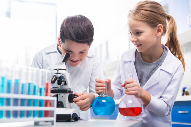 Genaue Arbeit. kräftige Junge Tuning Mikroskop, während schöne angenehme Mädchen ihm Lampen mit Flüssigkeiten gefüllt - Foto, Bild