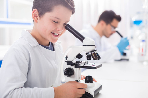 Take a look. Nice smart little boy touching microscope while looking in it and smiling - Фото, изображение