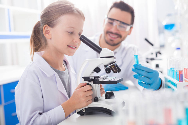 Work with microscope. Little smart beautiful girl smiling while working with microscope and dressing in lab coat - 写真・画像