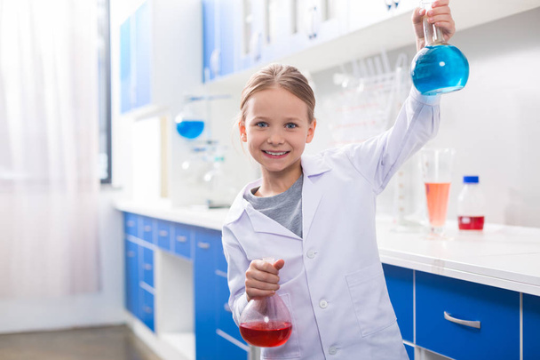Favourite science. Positive happy delighted girl smiling and holding flasks while being in the chemical lab - Фото, зображення