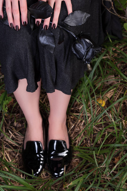 Γυναικεία πόδια σε μαύρο δερμάτινα υποδήματα ευρεσιτεχνίας στο γρασίδι. Μαύρη φούστα και ένα τριαντάφυλλο - Φωτογραφία, εικόνα