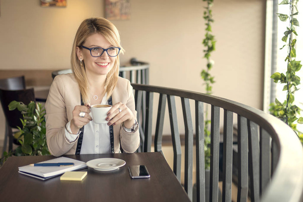 Joven mujer de negocios sonriente sentada en la cafetería, tomando café y pensando en algo
 - Foto, Imagen