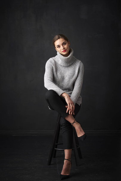 Full length shot of attractive young woman wearing turtleneck sweater while sitting at dark background and relaxing.  - Fotó, kép
