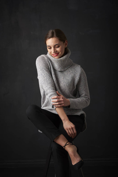Full length shot of attractive young woman wearing turtleneck sweater while sitting at dark background and relaxing.  - Φωτογραφία, εικόνα