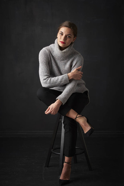 Full length shot of attractive young woman wearing turtleneck sweater while sitting at dark background and relaxing.  - 写真・画像