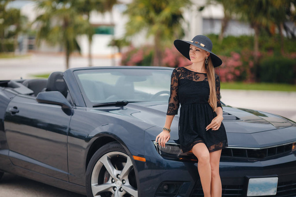 Young lady,brunette with long hair,beautiful young woman,wearing earrings in the ears is black summer dress sleeveless,head wears a big black hat,on the hand bracelet black,one travels through the resort area of America in a chic black convertible. - Фото, изображение