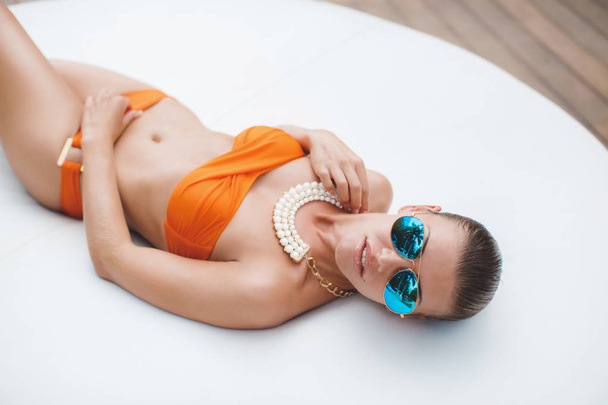 Elegant sexy woman in bikini on sun-tanned slim and shapely body posing near swimming pool. Gorgeous young woman posing in orange bikini lying by the pool in the mirror-blue glasses. - Fotoğraf, Görsel