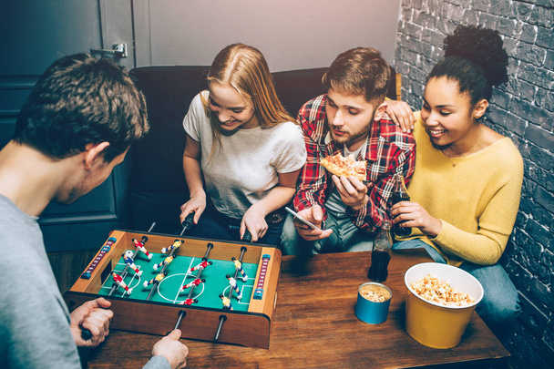Two people are playing football board game on the table while another two are watching on it and trying to support players as best as they can. Nobody knows who will win - Valokuva, kuva