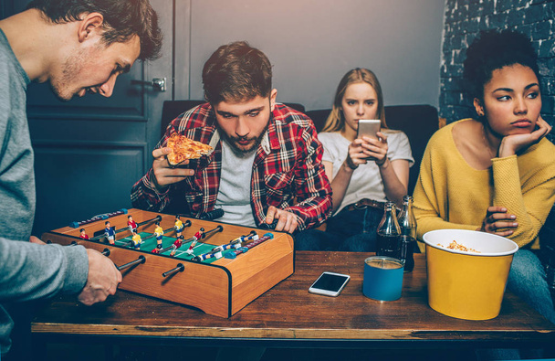 Two boys are playing board football game. They are very concentrated on it while girls are watching in the phone and feel boring. They dont like this game at all. - 写真・画像
