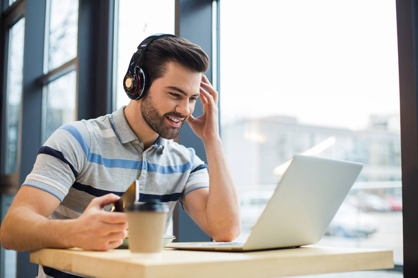 Αγαπημένο χόμπι. Όμορφος χαρούμενος ευτυχισμένος άντρας φορώντας ακουστικά και ακούγοντας μουσική ενώ κοιτάζοντας την οθόνη του φορητού υπολογιστή - Φωτογραφία, εικόνα
