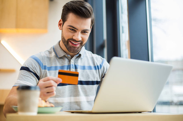 Electronic banking. Positive happy young man holding a credit card and smiling while making an online payment - Foto, Imagem