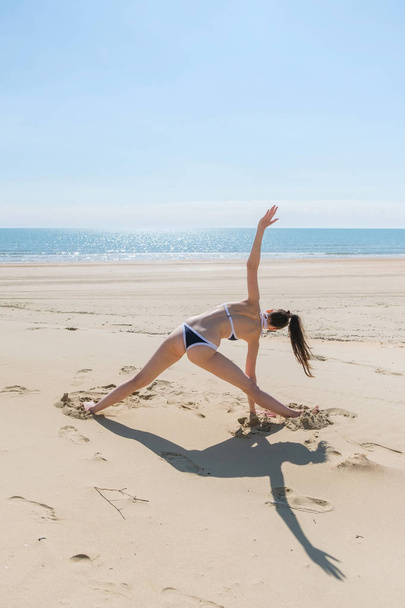 Unrecognizable woman in bikini doing yoga stands in a triangle position on the beach. - Fotó, kép